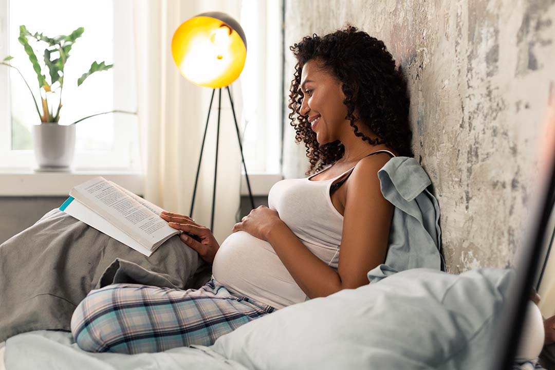 Pregnancy And Breast Implants - Pregnant woman resting on a bed while reading a book.
